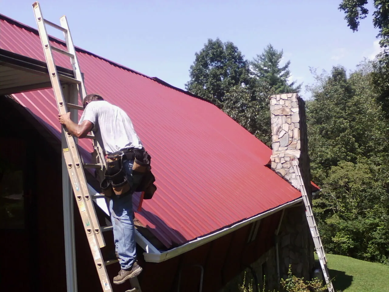 Roofer on a ladder beside a red metal roof and stone chimney — Soffit & Fascia Repair by Roofing Brothers in Mifflinburg